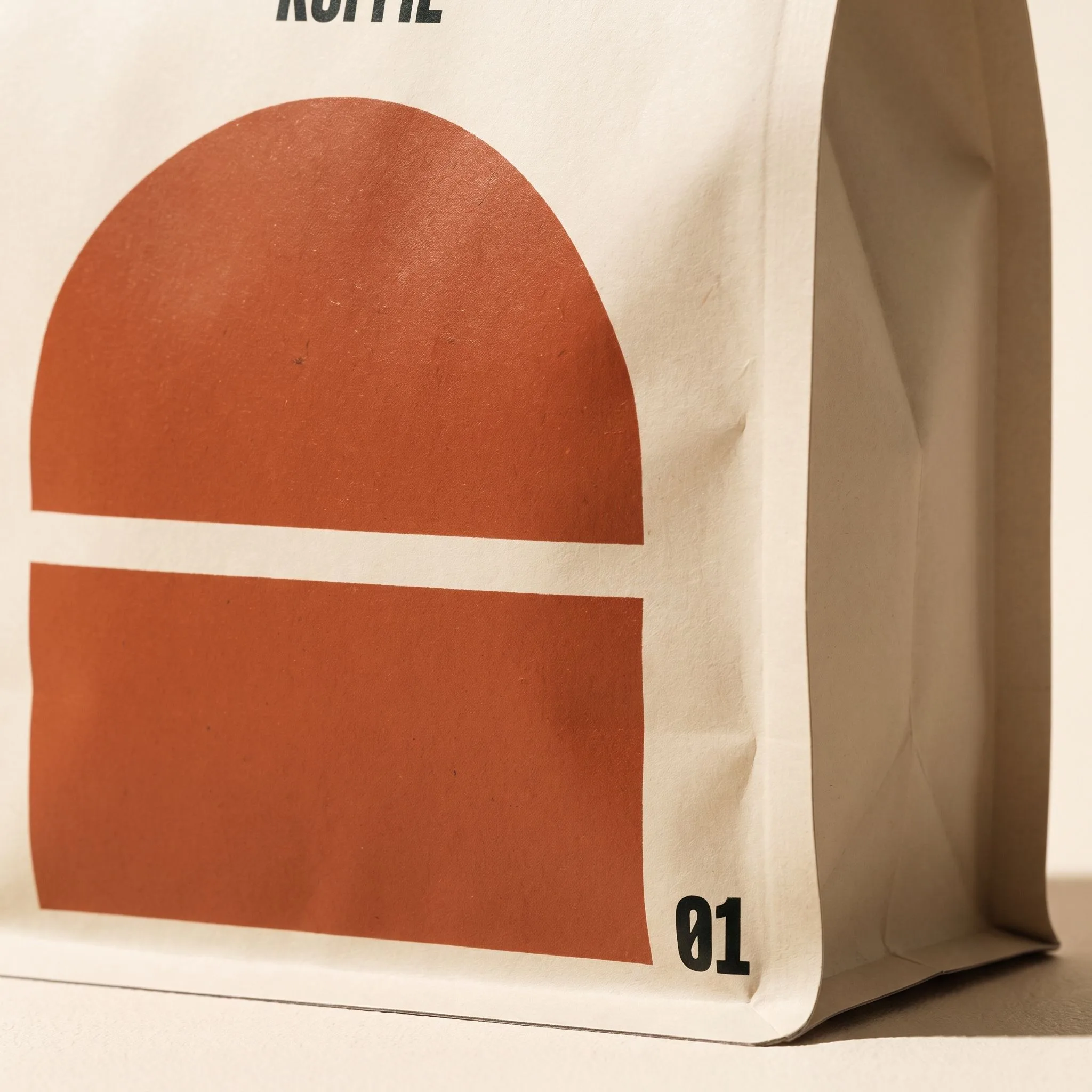 Detail van een roestoranje boog met een smalle crèmekleurige onderbreking op een matte KAAP-koffieverpakking.