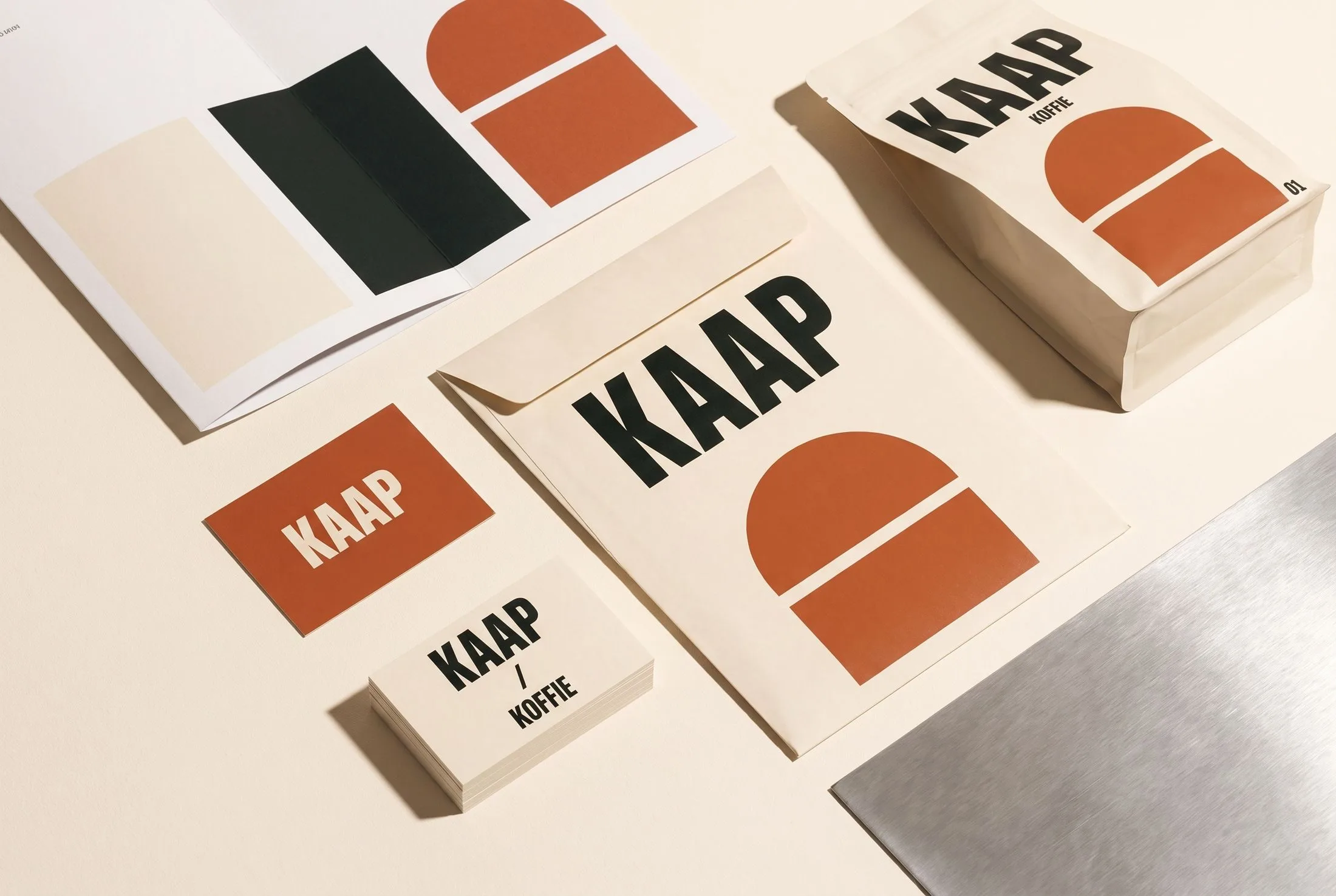 Bovenaanzicht van KAAP-drukwerk, kaarten, kleurvlakken en een koffieverpakking in crème, zwart en roestoranje.