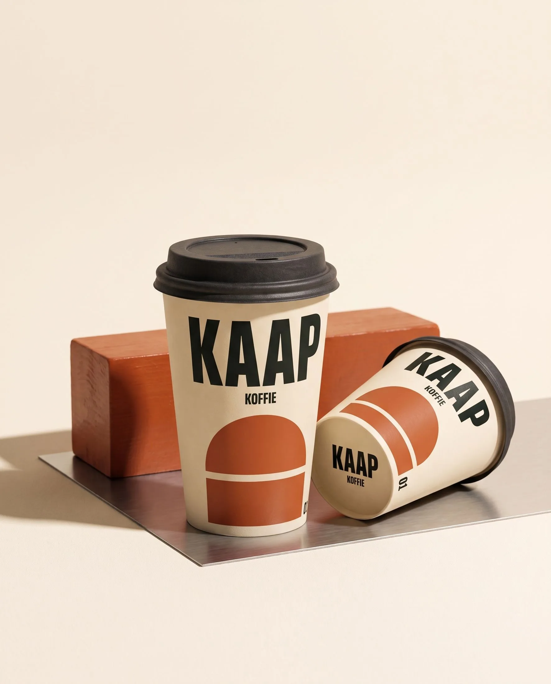 Twee crèmekleurige KAAP-koffiebekers met donkere deksels, naast een roestoranje blok.