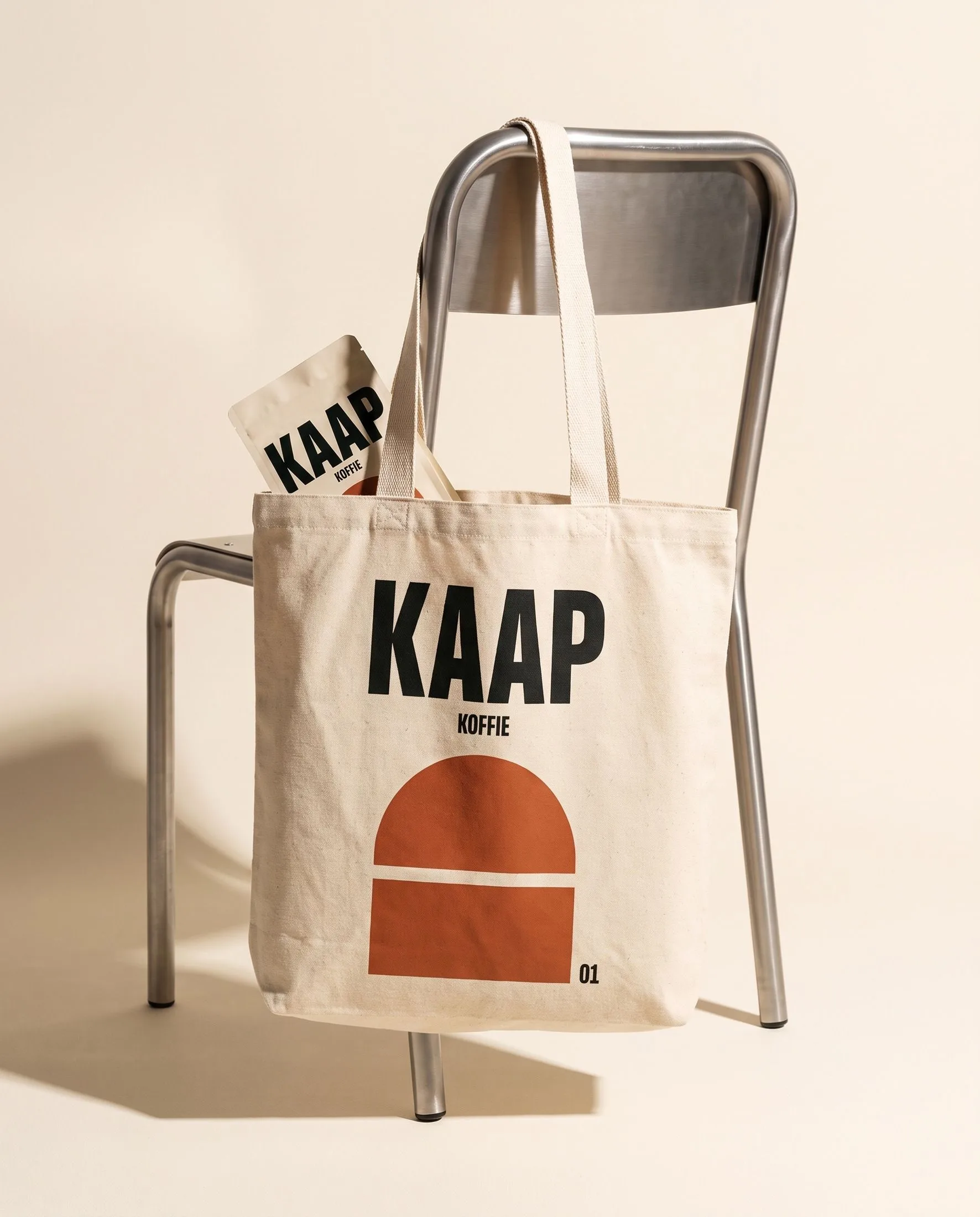 Een crèmekleurige canvas tas met het KAAP-woordmerk en een oranje boog hangt aan een metalen stoel.