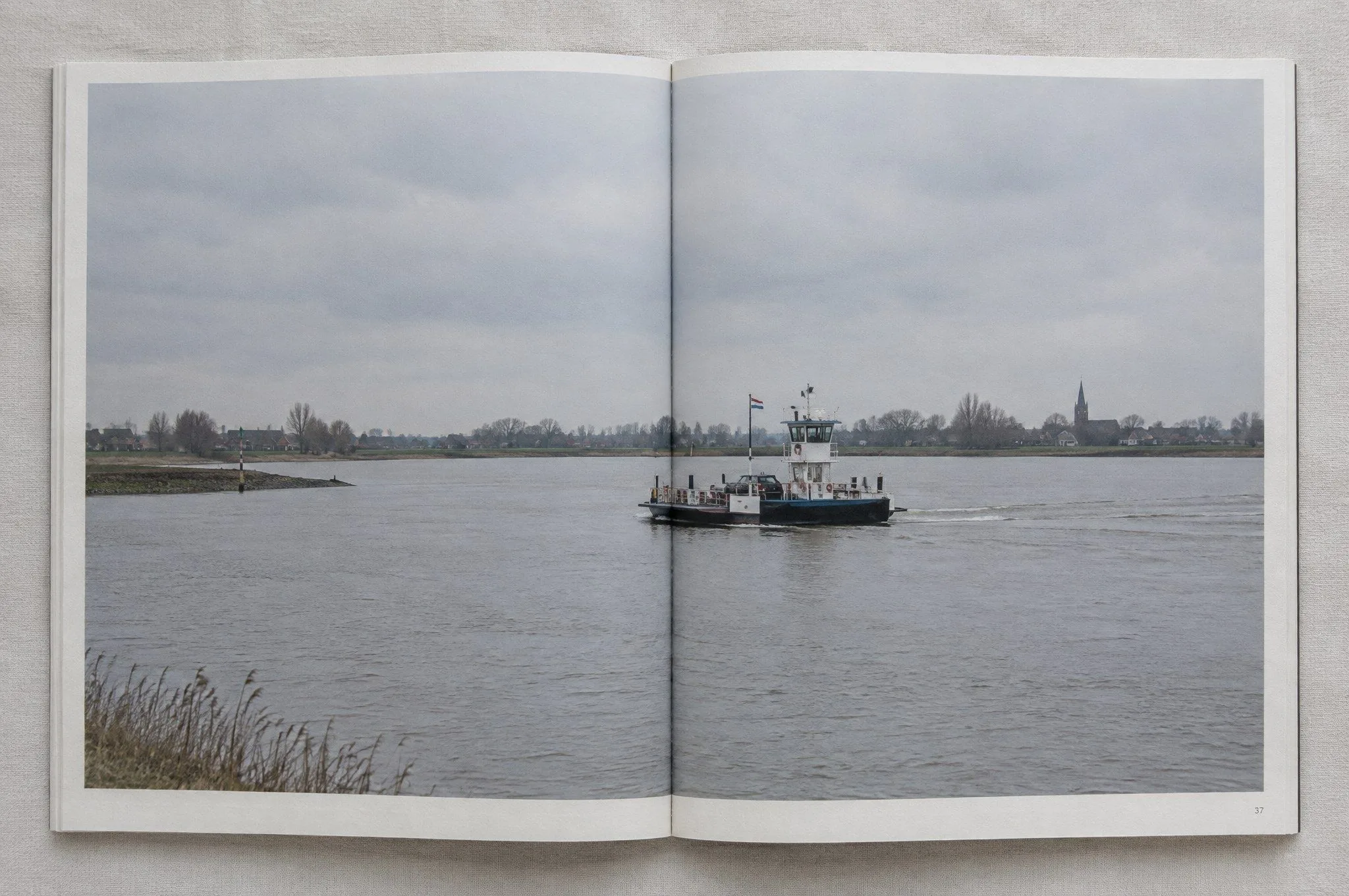 Boekspread met een foto over twee pagina’s van een kleine veerpont op een brede rivier.