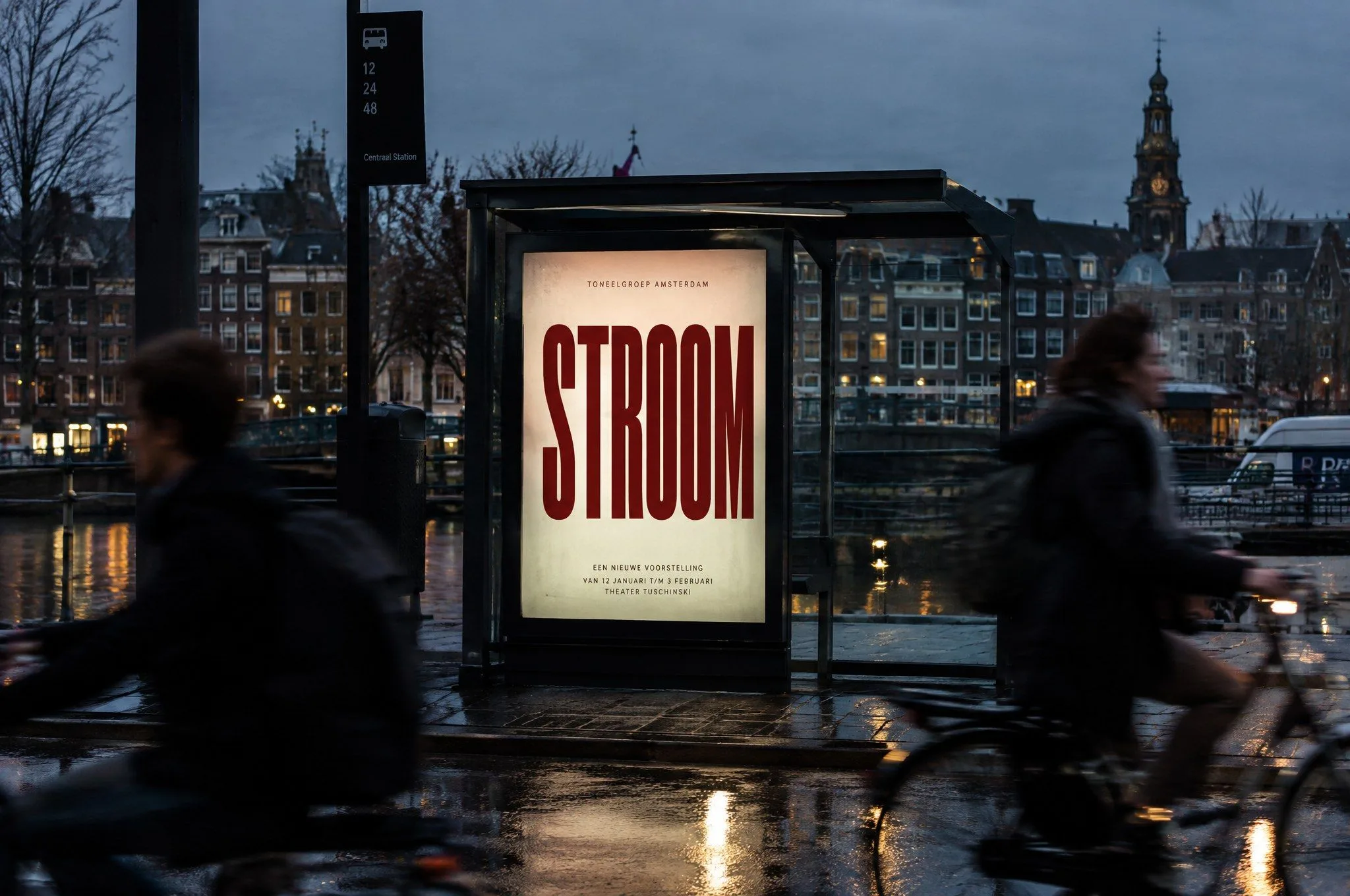 Een verlichte abri in de avond met het affiche STROOM, fietsers bewogen op de voorgrond.