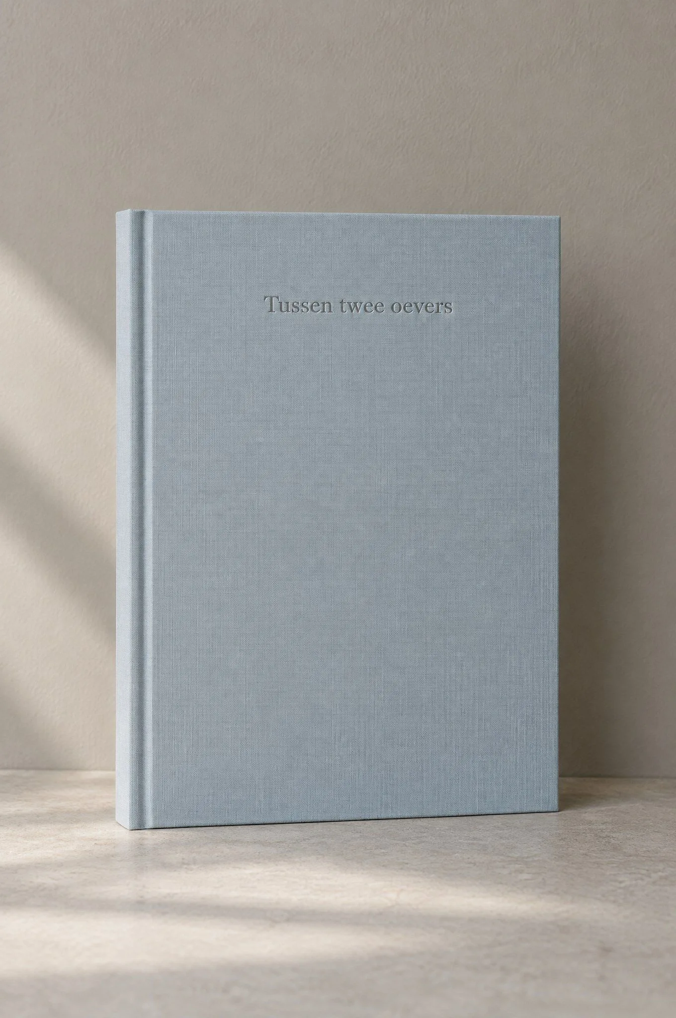 Een fotoboek met blauwgrijs linnen omslag en een blinddruk van de titel Tussen twee oevers.